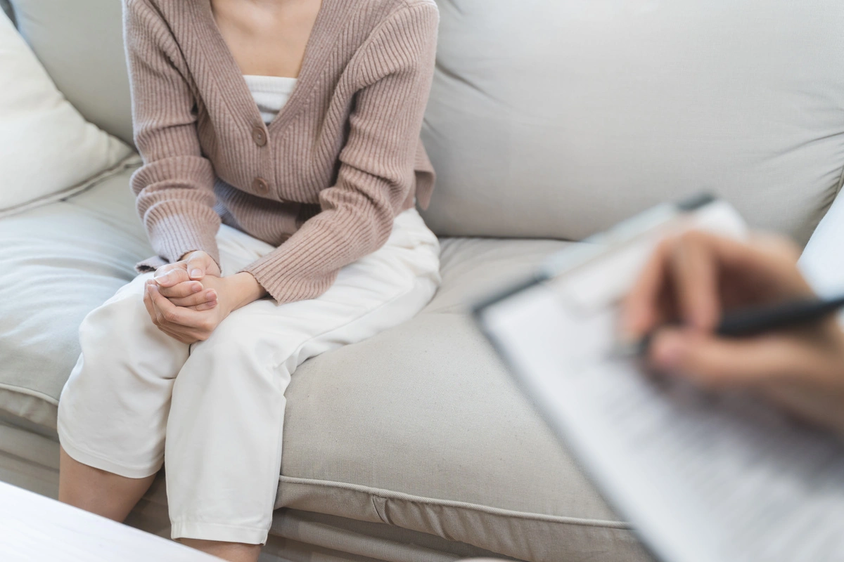 Une femme assise sur un canapé lors d'une consultation psy, les mains jointes, avec un thérapeute prenant des notes. Cette image montre une séance de thérapie individuelle.