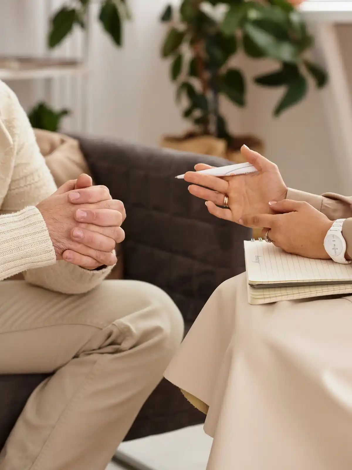 Un patient et un thérapeute en consultation. Le patient, vêtu d'un pull beige, est assis en face du thérapeute, les mains jointes. Le thérapeute, portant une robe beige, tient un carnet et un stylo, écoutant attentivement le patient tout en prenant des notes. L'environnement est apaisant avec des plantes en arrière-plan.