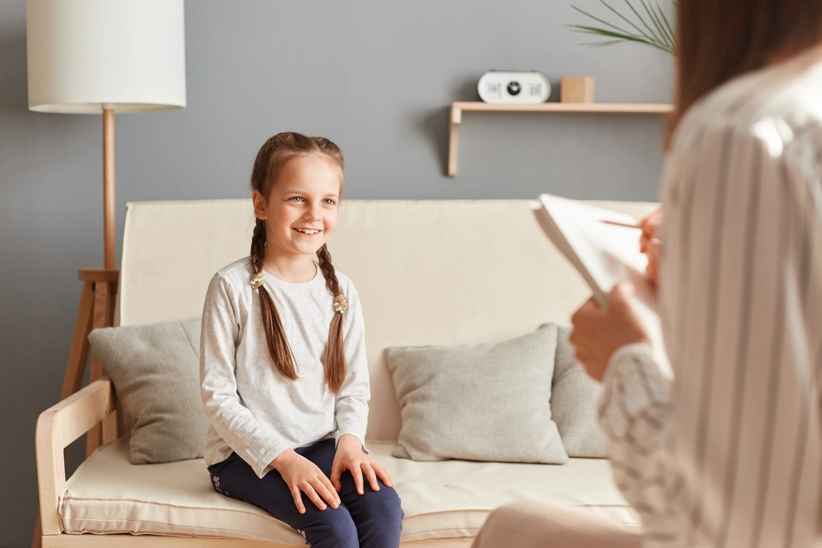 Image d'une thérapeute parlant avec une jeune fille souriante assise sur un canapé. La séance se déroule dans un environnement chaleureux et accueillant, démontrant l'approche bienveillante de la psychothérapie pour enfants, visant à aider les jeunes à surmonter leurs défis émotionnels et comportementaux.